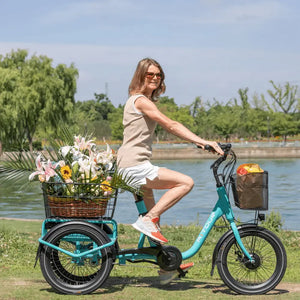 Woman riding the Aitour Heal Mini Folding Electric Trike 250W in blue near a river, with flowers and groceries in the baskets.