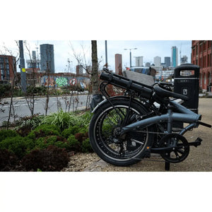 Folded Falcon Compact electric folding bike parked outdoors in an urban setting with city buildings in the background.