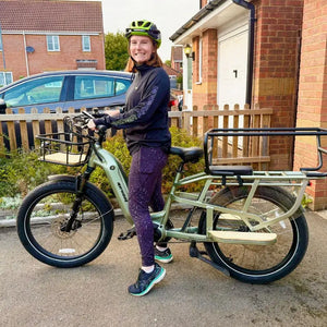A woman stands next to a light green Revom Multi C2 cargo e-bike with a large rear rack and front basket.