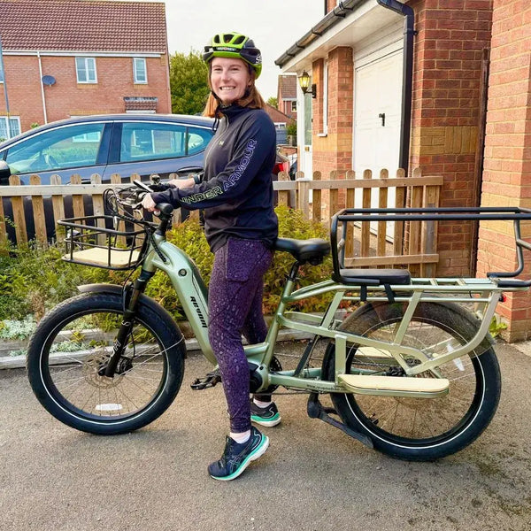 A woman stands next to a light green Revom Multi C2 cargo e-bike with a large rear rack and front basket.
