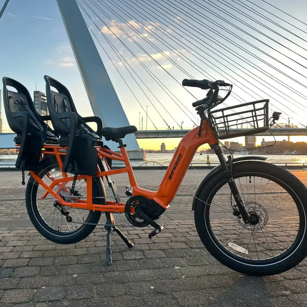 An orange Revom Multi C2 cargo e-bike with child seats and front basket, parked by a river in a city with a bridge in the background at sunset.