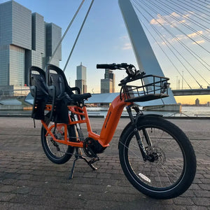 An orange Revom Multi C2 cargo e-bike with two seats and front basket, parked by a river in a city with a bridge in the background at sunset.
