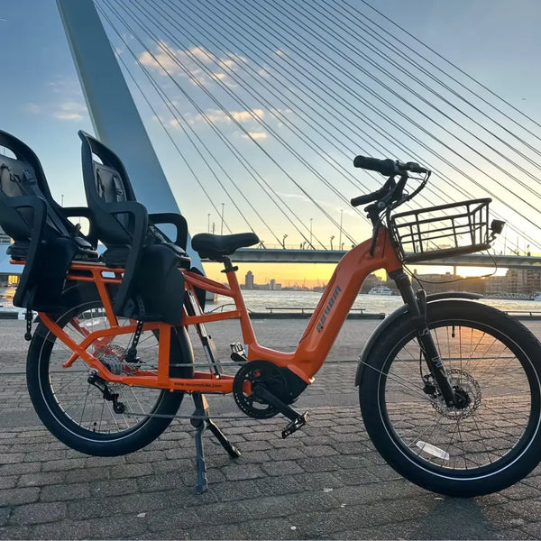 An orange Revom Multi C2 cargo e-bike with two seats and front basket, parked by a river in a city with a bridge in the background at sunset.