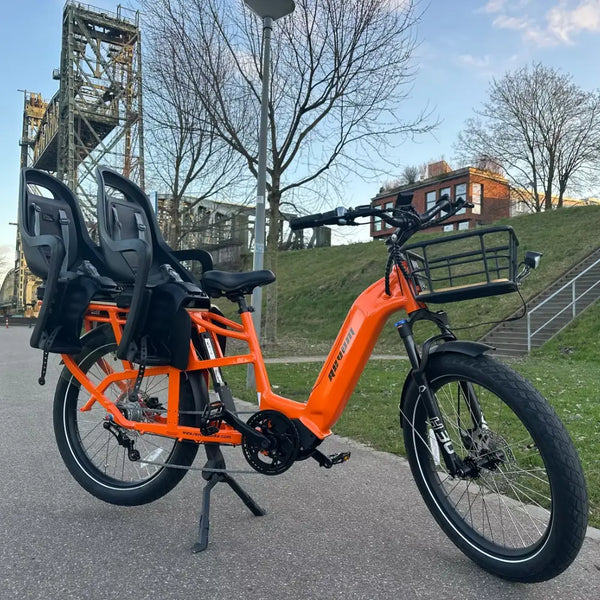An orange Revom Multi C2 cargo e-bike with two seats and front basket, parked on a paved path in an urban area.