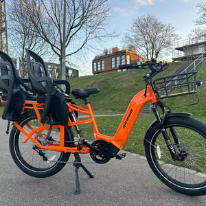 An orange Revom Multi C2 cargo e-bike with two seats and front basket, parked on a paved path in an urban area.