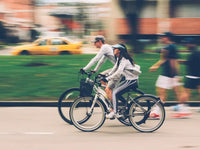 Two people riding electric bikes on a public road, showing where e-bikes can be used in urban areas including streets and cycle lanes.