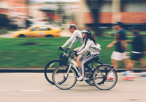 Two people riding electric bikes on a public road, showing where e-bikes can be used in urban areas including streets and cycle lanes.