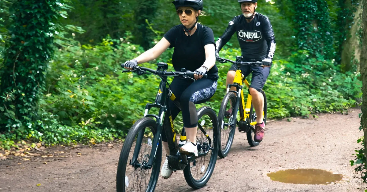 Man and woman riding electric mountain bikes together on a woodland trail.