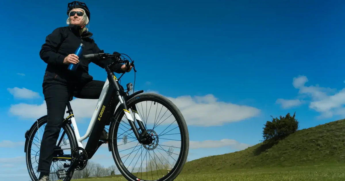 A woman riding a Mark2 X-Cross electric bike across open countryside under a bright blue sky.