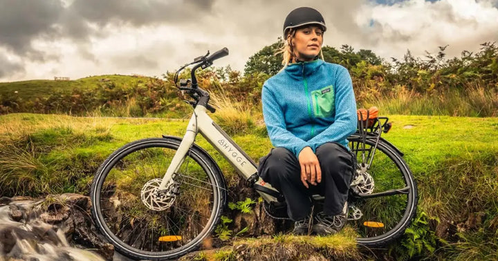 A woman wearing a helmet sits beside her electric bike in a scenic countryside landscape near a small stream.