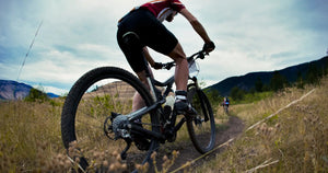 Cyclist riding an electric bike off-road on a mountain trail with grassy terrain and hills in the background.