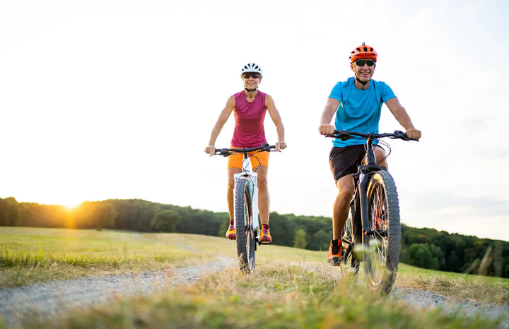 Man and woman riding electric bikes along a countryside trail at sunset with open fields in the background