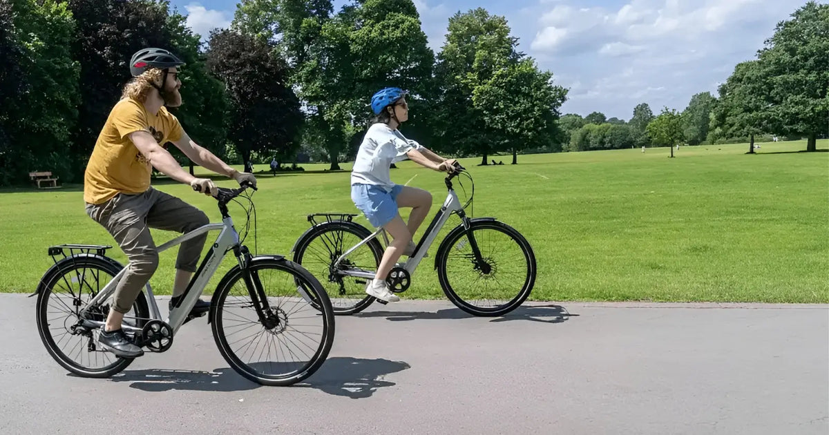 Two riders enjoying a relaxed ride on electric bikes through a sunny park - showing e-bikes as a practical alternative to cars for UK commuters
