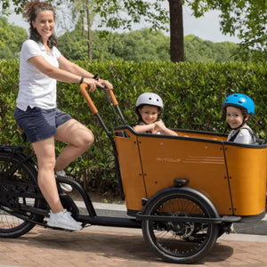 A mother riding the Aitour Family C electric cargo bike along a paved path with two happy children wearing helmets seated safely in the front wooden cargo box.