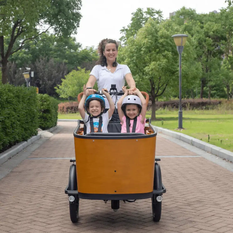 A mother riding the Aitour Family C electric cargo bike along a paved path with two happy children wearing helmets seated safely in the front wooden cargo box.
