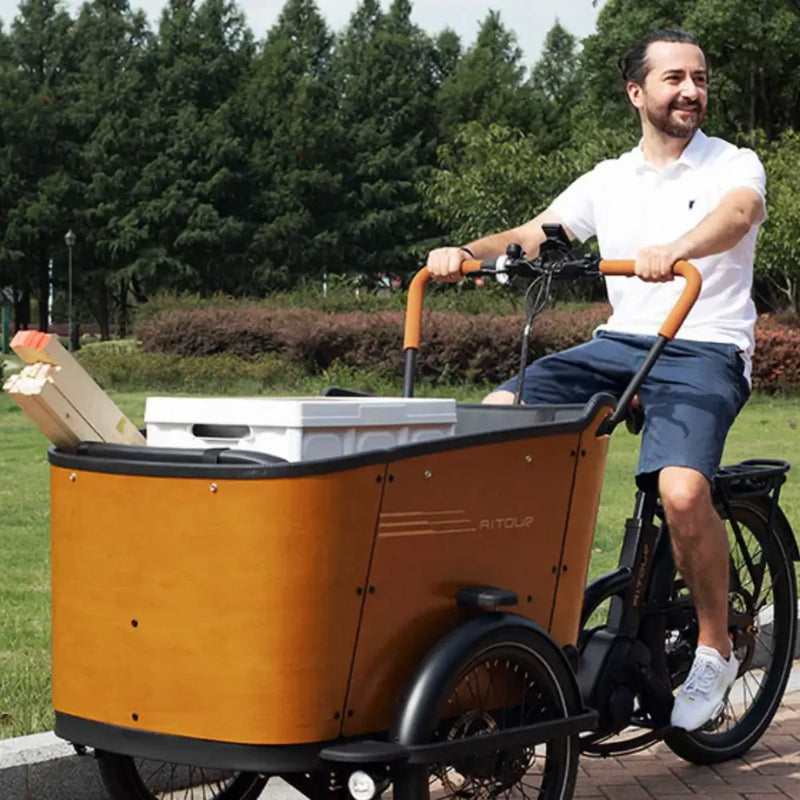 A man riding the Aitour Family C electric cargo trike through a park, demonstrating its capacity to transport goods and crates with ease in an outdoor setting.