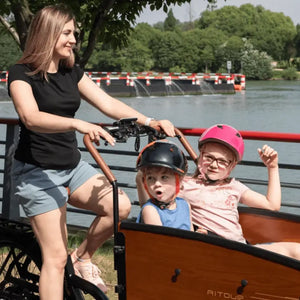 A woman riding the Aitour Family S electric cargo bike along a riverside path with two children wearing helmets seated safely in the front wooden cargo box.