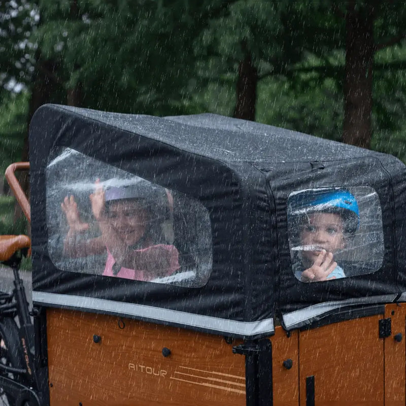 Lifestyle shot of two children sitting safely inside the Aitour Family S cargo box, protected from the rain by the weather-resistant canopy cover.