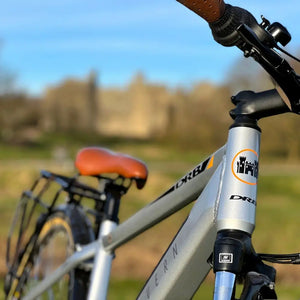 Close-up of the silver alloy frame and headtube on the Dallingridge Malvern electric bike, showing the weld details and branding against a sunny countryside backdrop.