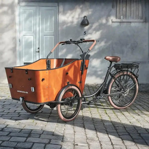 Front view of the Amcargobikes Ultimate Harmony electric cargo tricycle, featuring a spacious wooden cargo box, three-wheel stability, and brown puncture-resistant tyres on a cobblestone street.