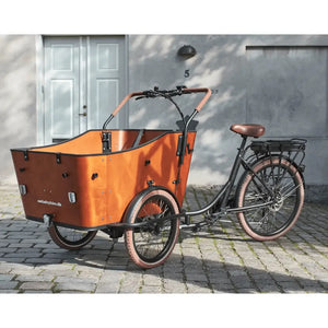 Front view of the Amcargobikes Ultimate Harmony electric cargo tricycle, featuring a spacious wooden cargo box, three-wheel stability, and brown puncture-resistant tyres on a cobblestone street.