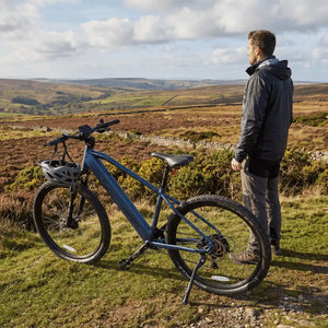 Man standing next to the Ampere X-Trail e-bike in a scenic landscape with rolling hills and a clear sky.
