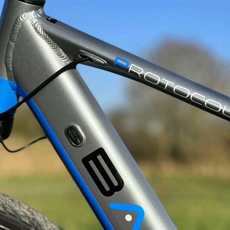 Close-up view of the Basis Protocol bike’s top tube featuring the “Protocol” logo and internal cable routing against a blue sky.