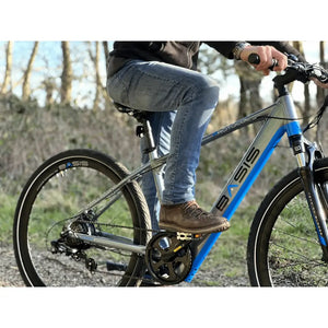 Rider sitting on the Basis Protocol electric hybrid mountain bike in an outdoor setting, showing the side profile and pedal position.