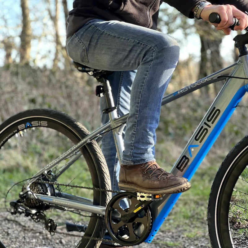Rider sitting on the Basis Protocol electric hybrid mountain bike in an outdoor setting, showing the side profile and pedal position.