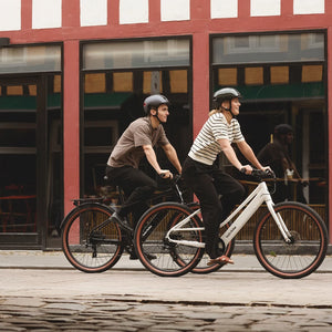 Two riders in helmets cycling a white Batribike Route S and a black Route X past a red timber building