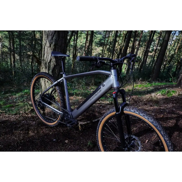 Angled view of the Claud Butler Ridge 2.0 Electric Mountain Bike (2025) in a forest, highlighting its suspension fork, rugged design, and tan wall tyres for trail riding.