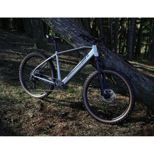 Side view of the Claud Butler Ridge 2.0 Electric Mountain Bike (2025) leaning against a tree in the woods, showcasing its off-road-ready design, suspension fork, and knobby tyres.