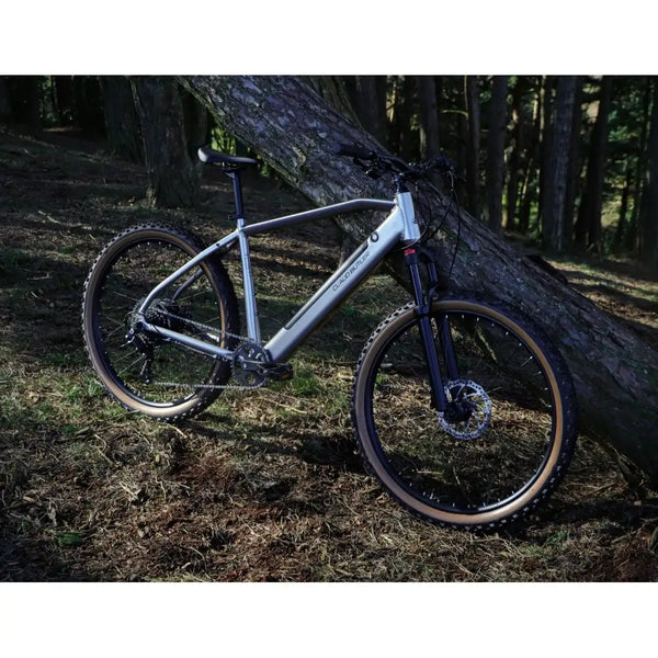 Side view of the Claud Butler Ridge 2.0 Electric Mountain Bike (2025) leaning against a tree in the woods, showcasing its off-road-ready design, suspension fork, and knobby tyres.