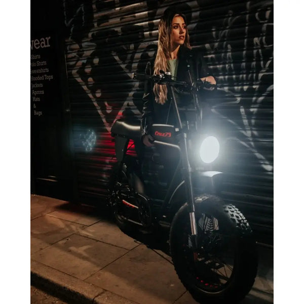 Woman standing beside the Cruz73 Pro Retro Fat Tyre Electric Bike at night in an urban setting with graffiti background and headlight on.