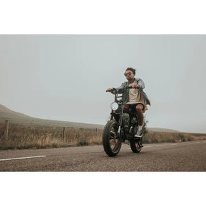 Man riding the Cruz73 Pro Retro Fat Tyre Electric Bike on an open countryside road, wearing sunglasses and casual clothing.