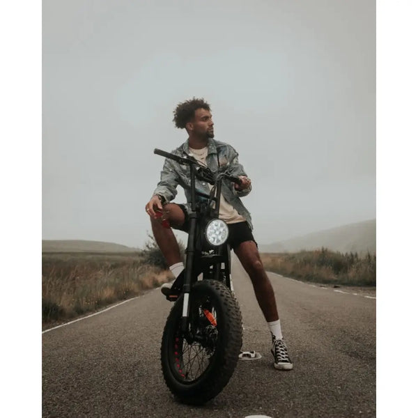 Man sitting on the Cruz73 Pro Retro Fat Tyre Electric Bike in the middle of a countryside road, dressed in a denim jacket and casual shorts.