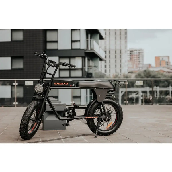 Cruz73 Retro Fat Tyre Electric Bike parked in an urban location with buildings in the background, showcasing its sleek design.