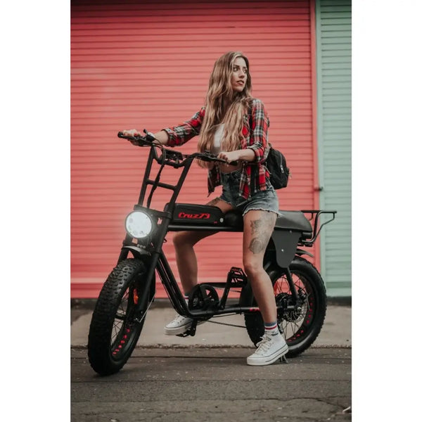 Woman in casual clothing sitting on a Cruz73 Retro Fat Tyre Electric Bike in front of a colourful shutter background, ready to ride.
