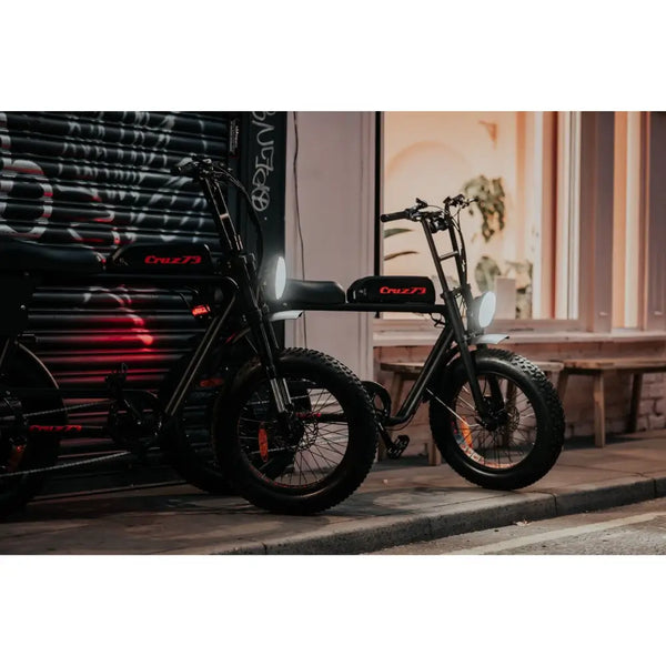 Two Cruz73 Retro Fat Tyre Electric Bikes with headlights on, parked along a dimly lit city street at night.