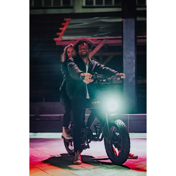 Two riders on the Cruz73 Unlimited Retro Fat Tyre Electric Bike at night, illuminated by the bright headlight in an urban setting.