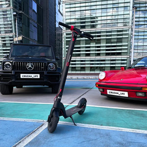 The black Cruzaa Commuta 350W electric scooter displayed alongside a Mercedes G-Wagon and Porsche 911 in a modern city setting.