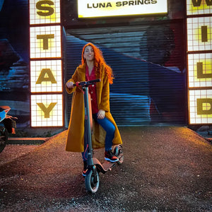 A stylish woman posing with the Cruzaa Commuta 350W electric scooter at night in front of a neon 'Stay Wild' sign.