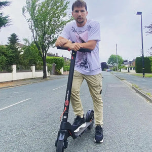 A rider in a black outfit with tattoos standing next to the Cruzaa Commuta 350W electric scooter on a residential street.