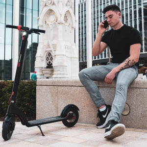 A commuter taking a break in a city plaza, sitting on a stone wall next to his parked Cruzaa Commuta electric scooter.