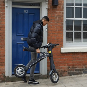 A man lifting the folded black Cruzaa E-Scooter Pro outside a residential home with a blue door, demonstrating its lightweight design and portability.
