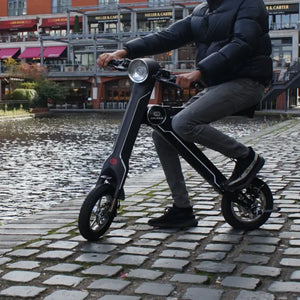 A man in a black puffer jacket riding the black Cruzaa E-Scooter Pro along a cobbled canal path, showcasing its versatility on different urban surfaces.