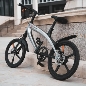 Angled front view of the grey Cruzaa e-bike leaning against a stone wall, highlighting the front suspension fork and perforated disc brake rotor.