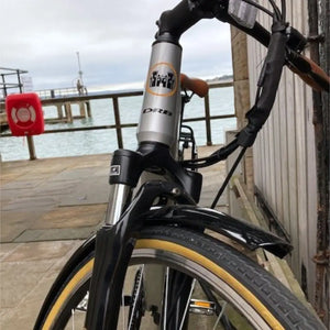 Front detail of the Dallingridge Malvern electric bike showing the headtube logo badge and suspension fork with a coastal pier and water in the background.