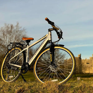 Angled front view of the Dallingridge Malvern hybrid electric bike highlighting the SR Suntour suspension fork and tan-wall tyres with a historic castle in the background.
