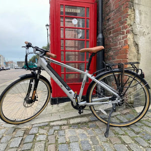 Side profile of the silver Dallingridge Malvern electric hybrid bike parked next to a classic red telephone booth on a cobblestone street.